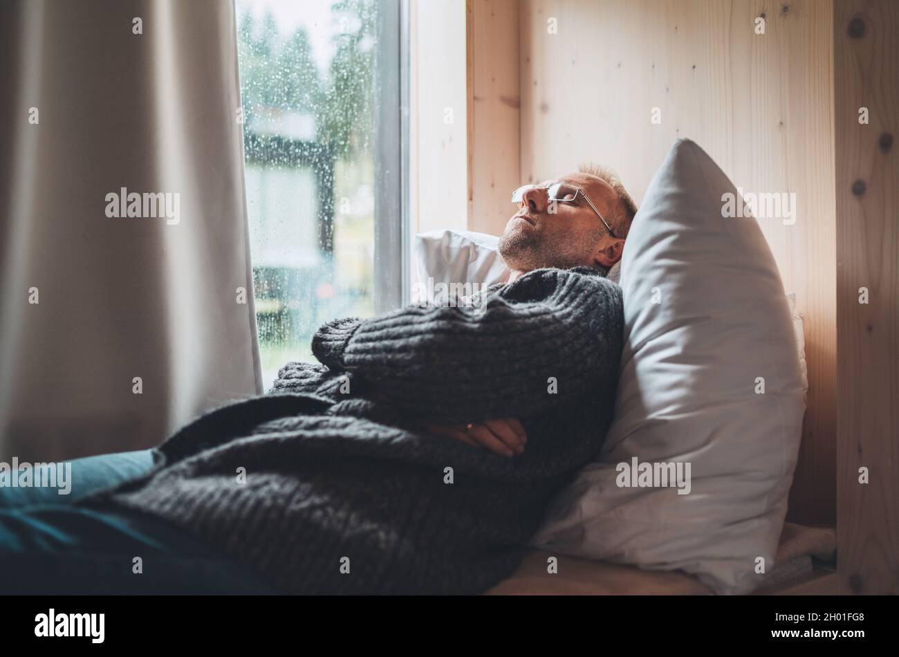 Dormant homme d'âge moyen en lunettes vêtu d'un gilet ouvert allongé sur un oreiller confortable à côté de la fenêtre.Image de concept de santé mentale et de temps d'automne. Banque D'Images