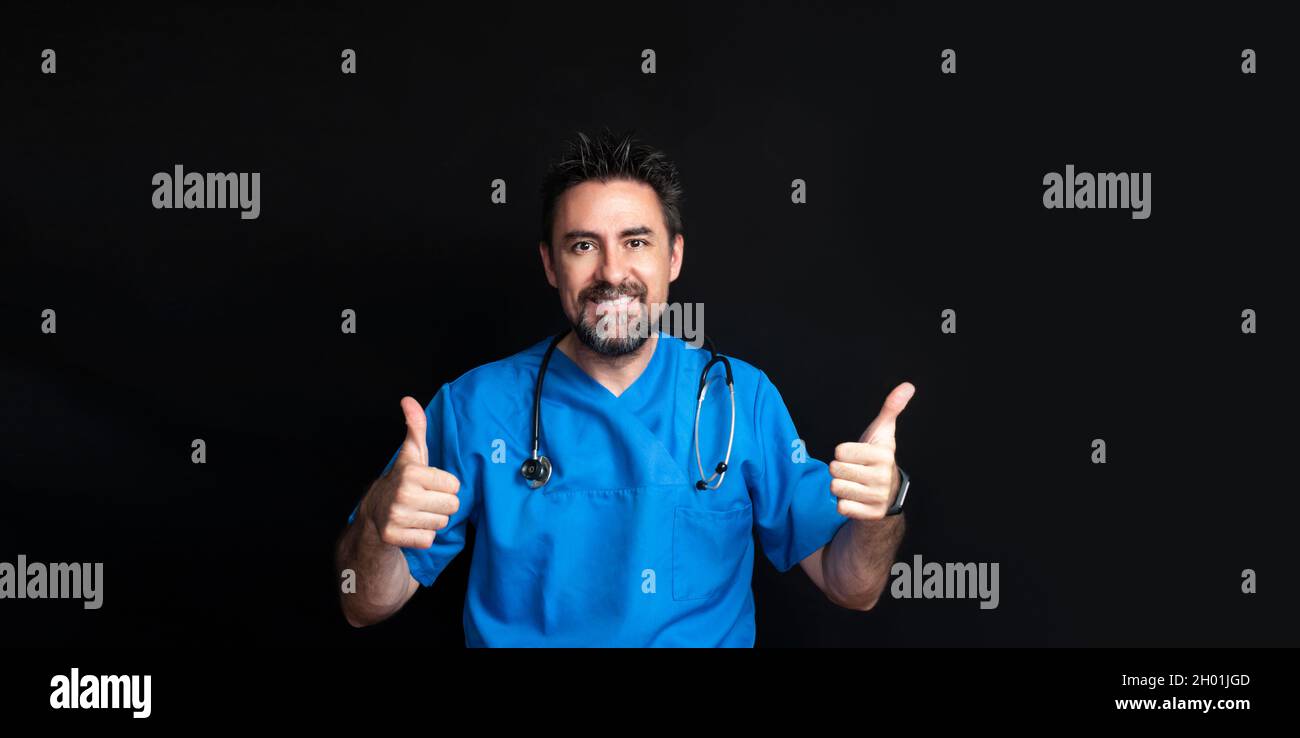 Un médecin vêtu d'un uniforme bleu d'hôpital, sombre et barbu, avec un stéthoscope autour de son cou, souriant et levant ses deux pouces vers le haut pour indiquer le Th Banque D'Images