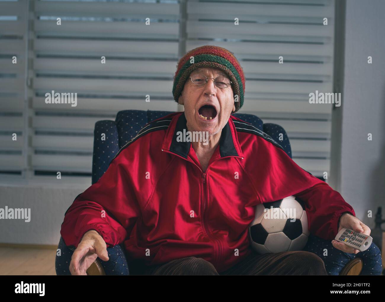 Un homme senior enthousiaste assis dans un fauteuil à la maison, tenant le ballon de football et regardant un match de football à la télévision Banque D'Images