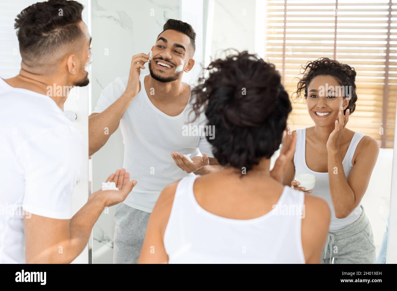 Routine du matin.Happy Young Arab couple se préparer ensemble près du miroir dans la salle de bains, femme appliquant la crème hydratante tandis que l'homme utilisant la mousse de rasage, Banque D'Images