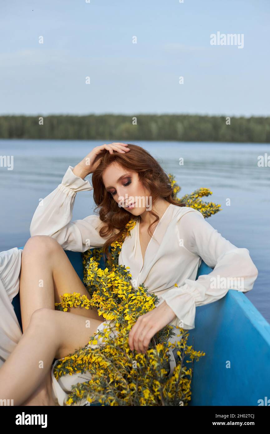 REDHEAD femme en robe de négligence blanche s'asseoir dans le bateau bleu sur le lac d'étang. Fille à cheveux rouges avec branches de fleur jaunes assis dans le bateau au coucher du soleil Banque D'Images