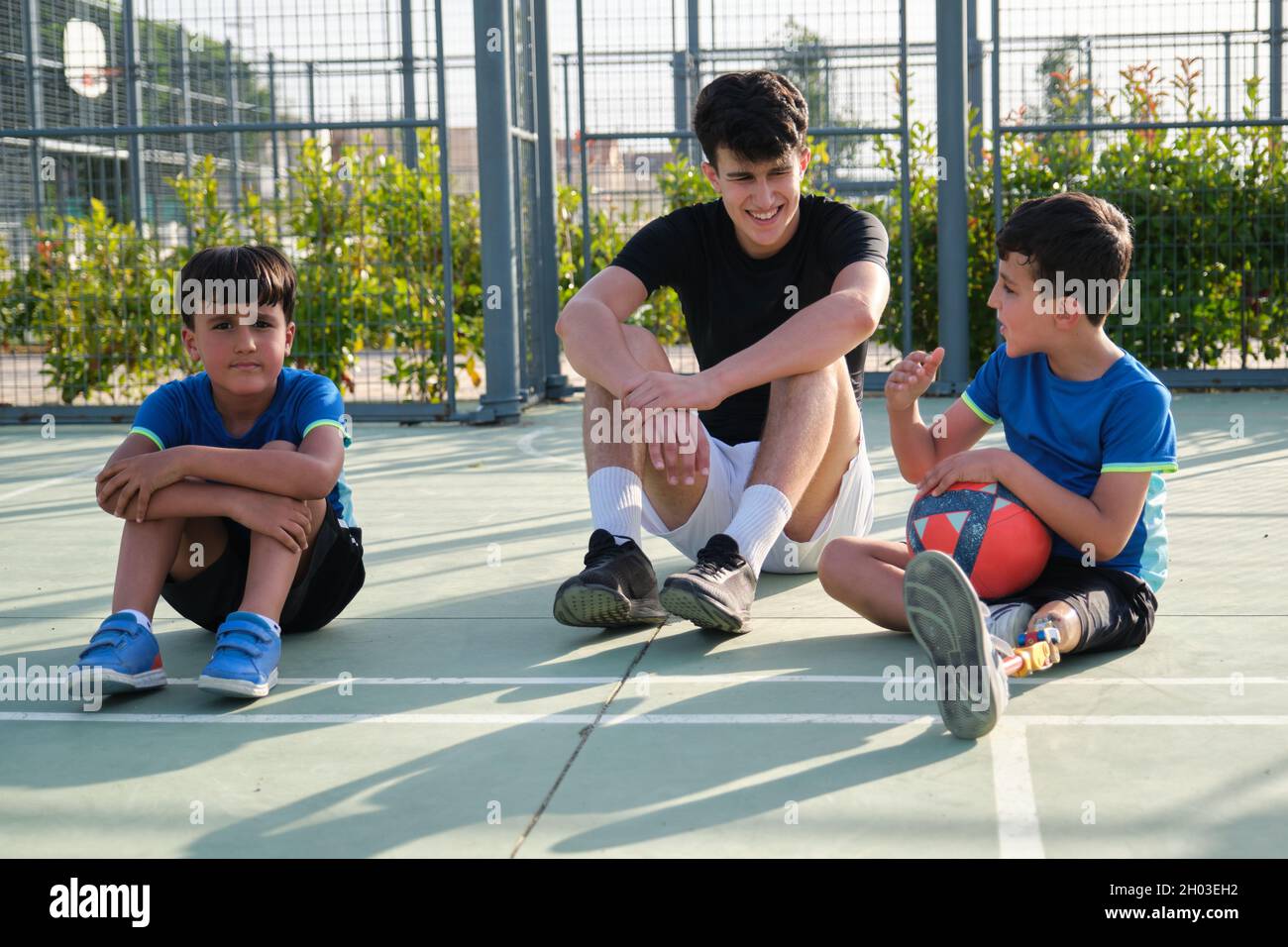 Un entraîneur s'assoit au sol du terrain de football pour se reposer et parler avec deux élèves. Banque D'Images