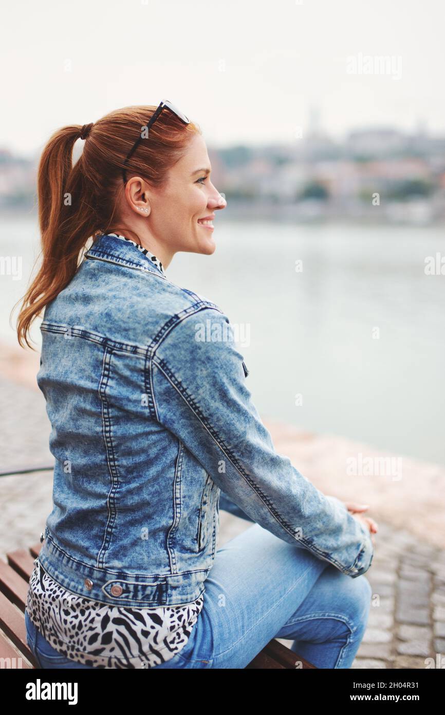 Bonne jeune femme caucasienne urbaine à tête rouge regardant loin au bord de la rivière, vue de profil Banque D'Images