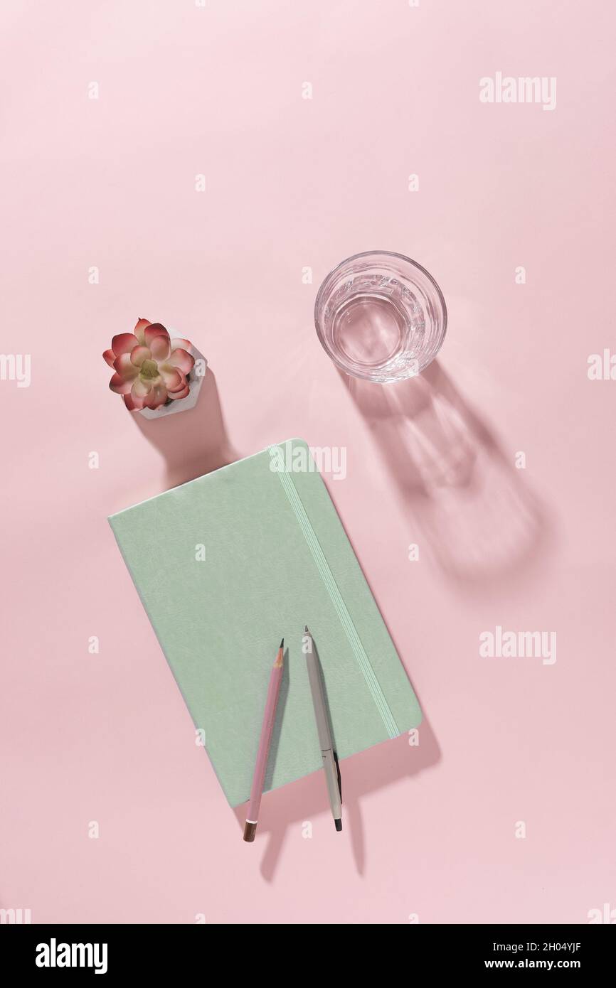 Arrière-plan de bureau rose pastel avec carnet vert fermé, verre d'eau douce avec ombre, fleur et stylo dans un journal d'affaires, de créativité ou personnel Banque D'Images