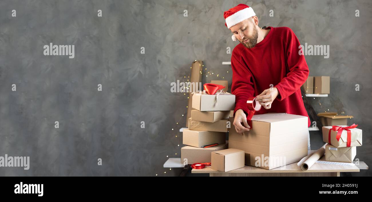 Le propriétaire d'une petite entreprise est un entrepreneur mâle barbu qui fait l'emballage de marchandises à la maison dans un petit bureau.Prépare les boîtes pour l'expédition.Grande vente pour Noël.Baner. Banque D'Images