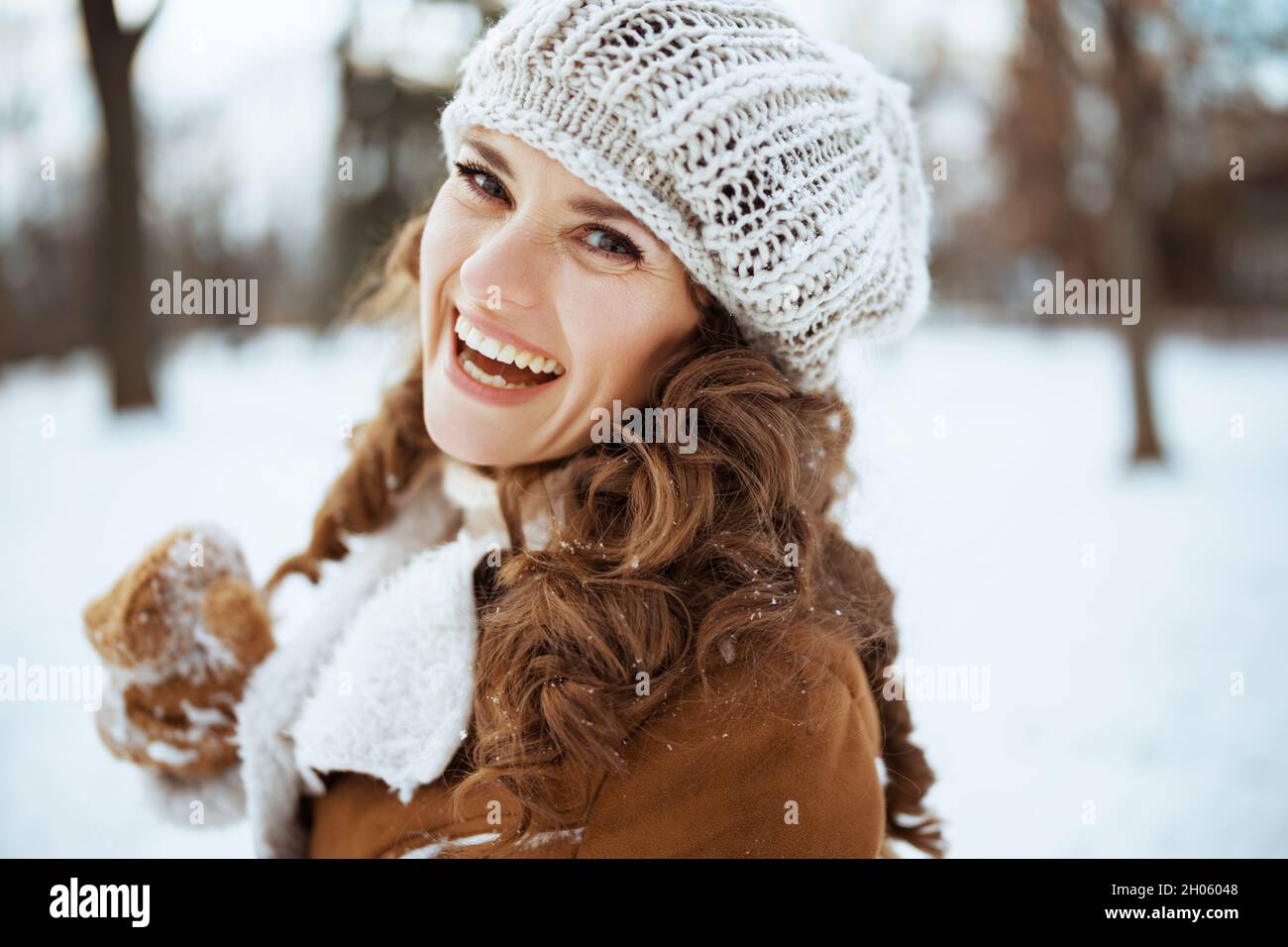 Portrait d'une femme heureuse et moderne de 40 ans avec des moufles dans un  bonnet tricoté et un manteau de peau de mouton dehors dans le parc de la  ville en hiver