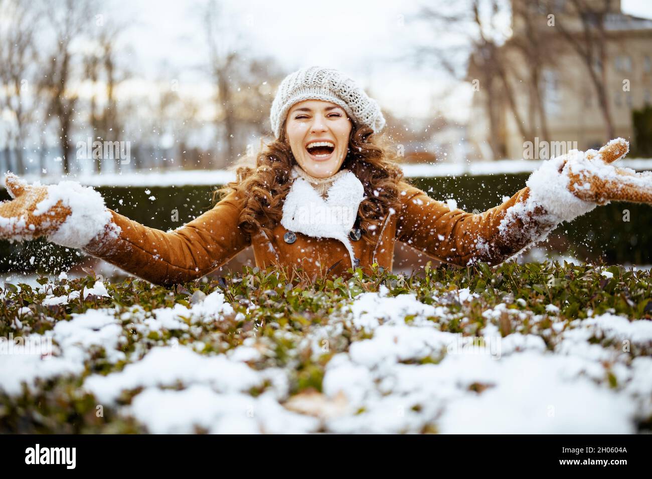 femme élégante et souriante avec moufles dans un bonnet tricoté et un  manteau en peau de mouton jetant de la neige à l'extérieur dans le parc de  la ville en hiver Photo
