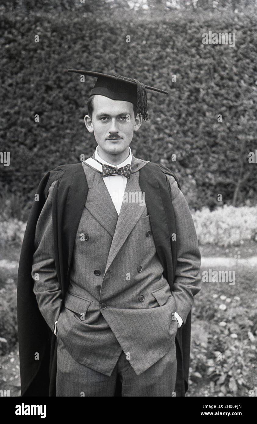 Années 1950, historique, un jeune anglais avec une courte moustache et  portant un costume double et un noeud papillon posant pour sa photo dans  une robe de graduation et une casquette universitaire