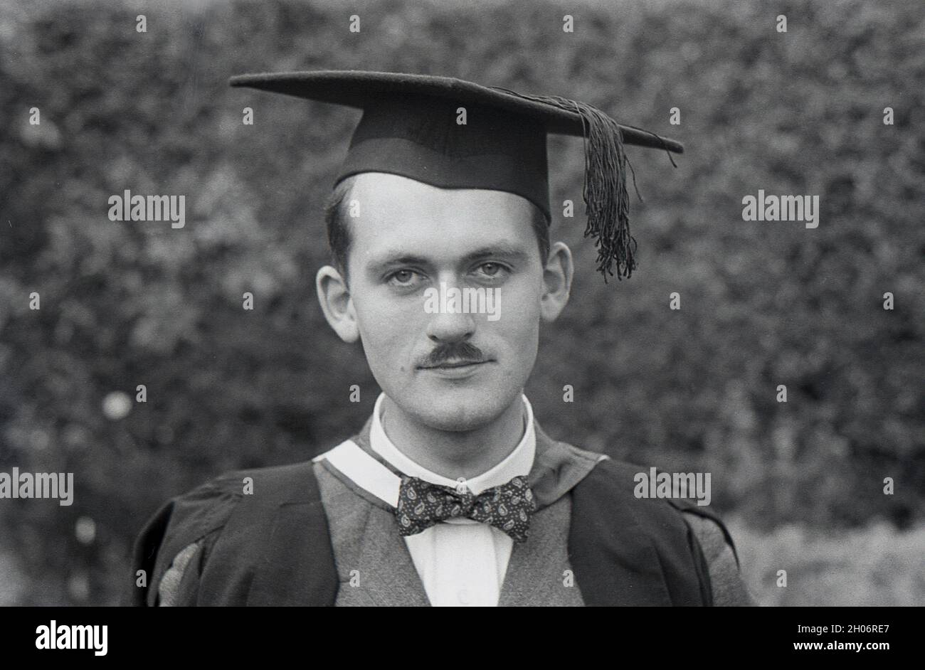 Années 1950, photo historique en gros plan d'un jeune homme avec une courte  moustache et portant un costume double et un noeud papillon posant pour son  image dans une robe de graduation