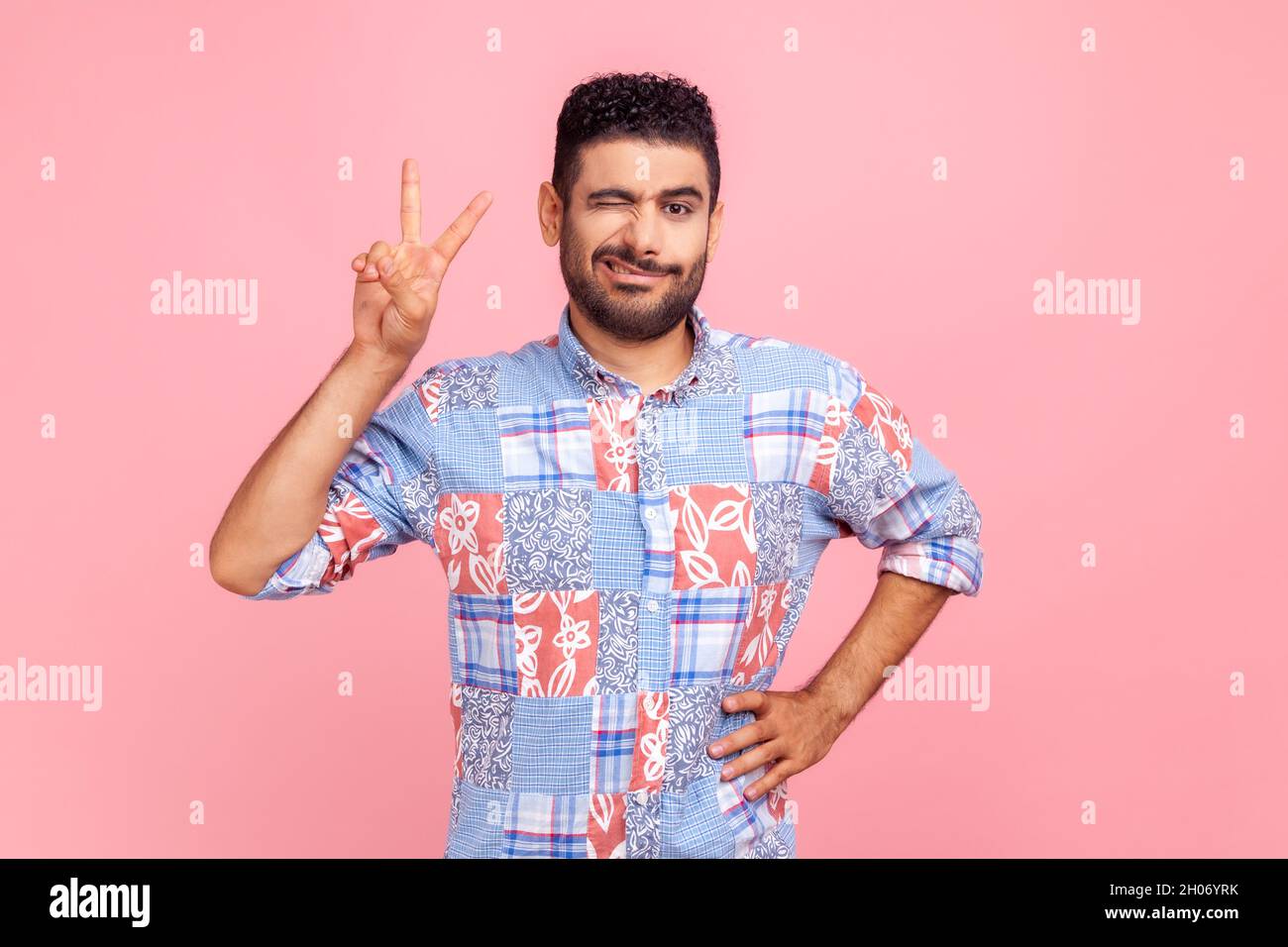 Portrait de l'homme excité qui se fait un clin d'œil et qui frognait le visage dans une chemise bleue décontractée debout avec main levée et montrant le signe v ou la paix, le geste de victoire.Studio d'intérieur isolé sur fond rose. Banque D'Images