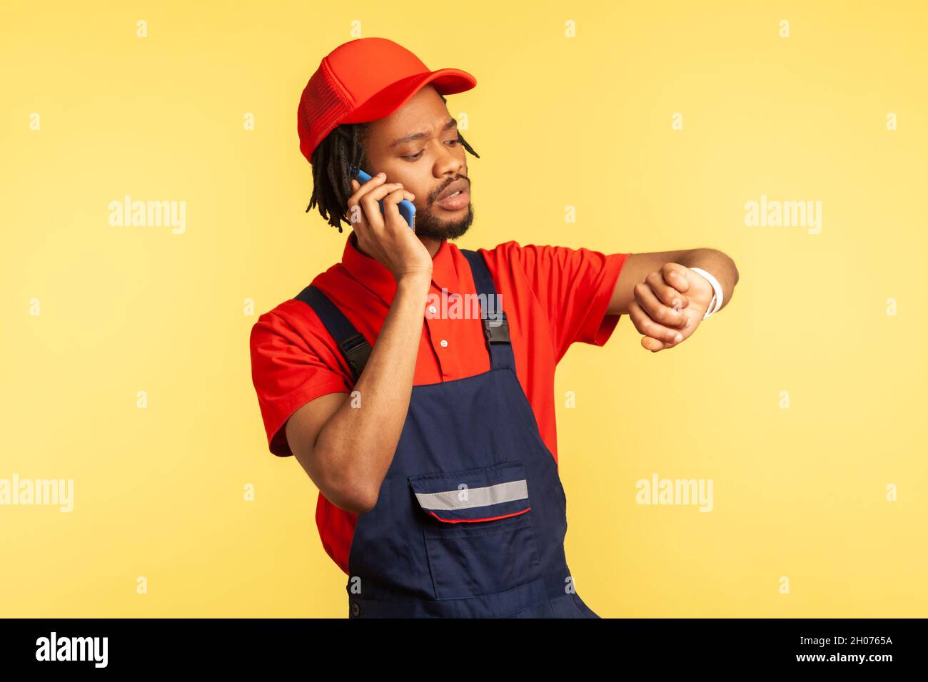 Portrait d'un coursier concentré portant une combinaison bleue parlant téléphone avec le client, regardant la montre intelligente, service de livraison à temps.Studio d'intérieur isolé sur fond jaune. Banque D'Images