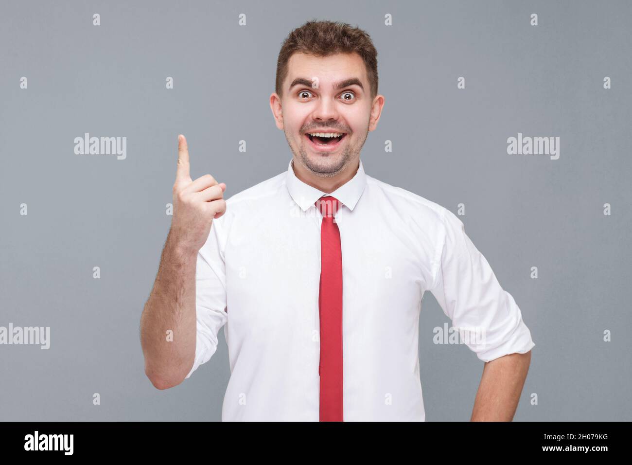 Idée.Portrait d'un jeune beau homme surpris en chemise blanche et cravate debout, le doigt vers le haut et regardant l'appareil photo avec l'idée geste et profiter. Intérieur isolé sur fond gris. Banque D'Images