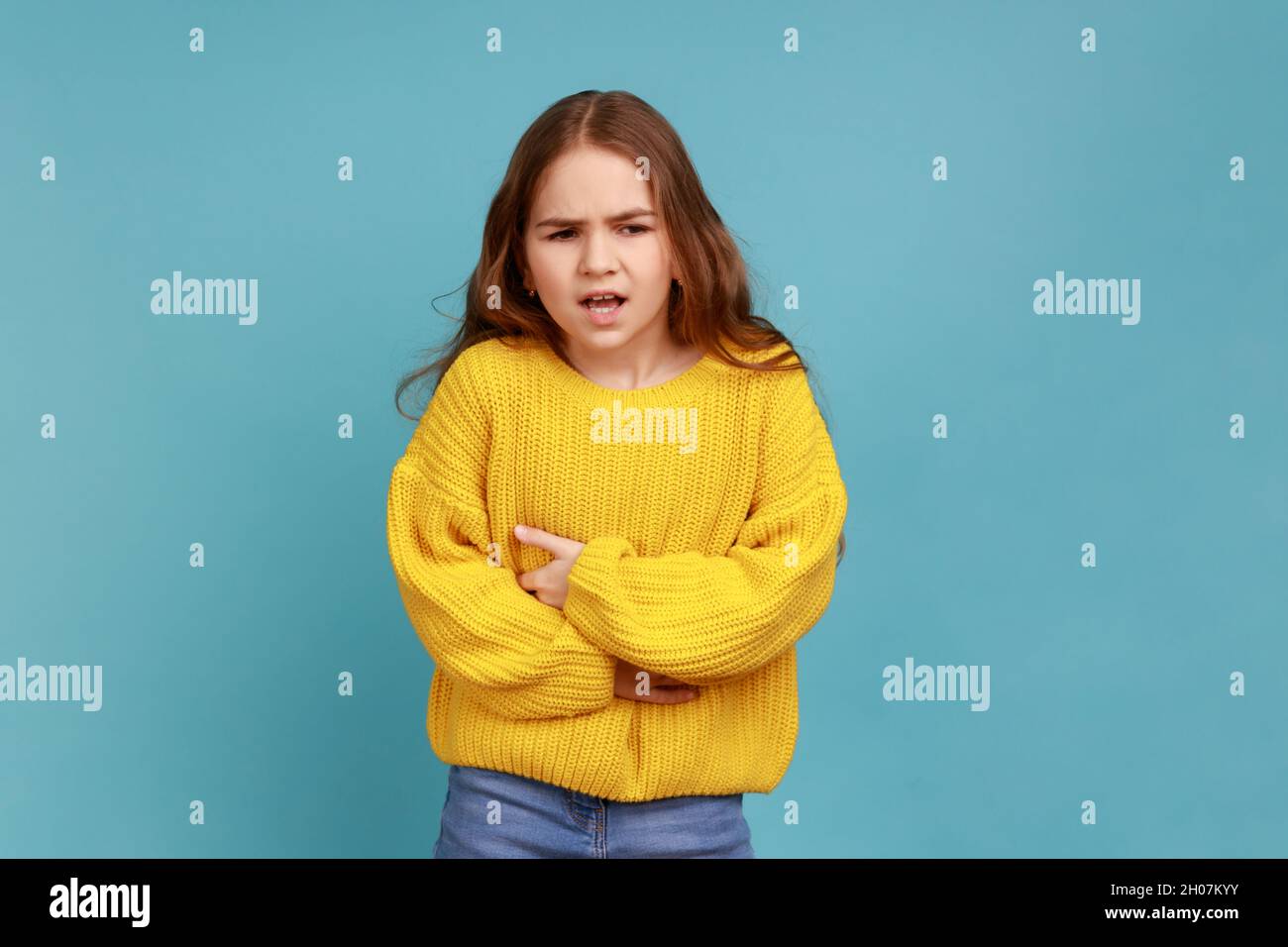 Petite fille embrassant le ventre, sent l'inconfort ou la douleur dans l'estomac, souffrant des crampes de constipation, portant le chandail jaune de style décontracté.Studio d'intérieur isolé sur fond bleu. Banque D'Images