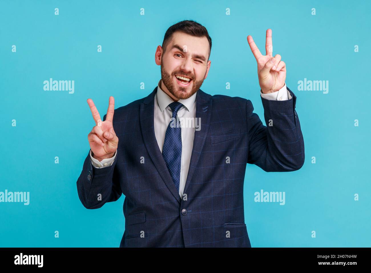 Joyeux homme d'affaires positif portant le costume officiel de style montrant le signe v à l'appareil photo, en se clin d'œil et souriant, jeune homme souhaitant bonne chance, paix.Studio d'intérieur isolé sur fond bleu. Banque D'Images