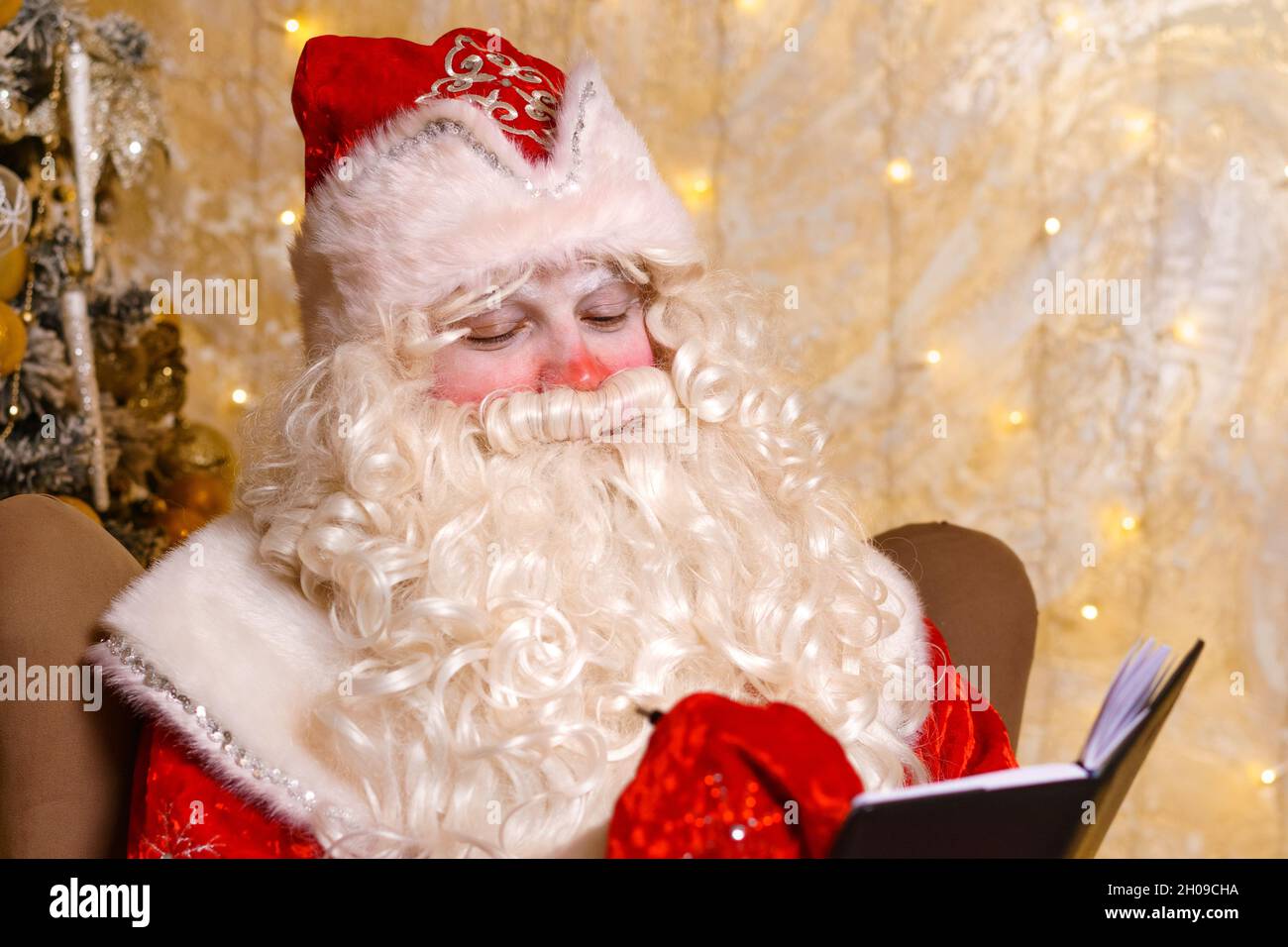 Le Père noël est assis sur une chaise, contre le fond des écrits de l'arbre de noël dans le carnet.Regarde la liste des cadeaux de noël pour les enfants.Réalisation des désirs à la veille du nouvel an. Banque D'Images