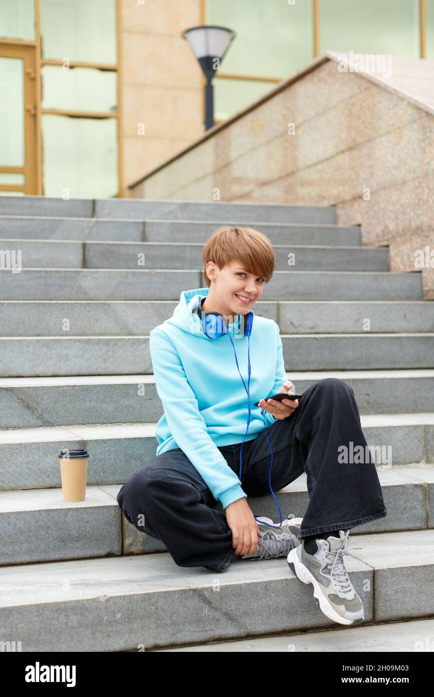 Jeune femme blogger est assis sur les escaliers avec un smartphone.Elle sourit et regarde l'appareil photo.Photo verticale.Concept de technologie moderne. Banque D'Images