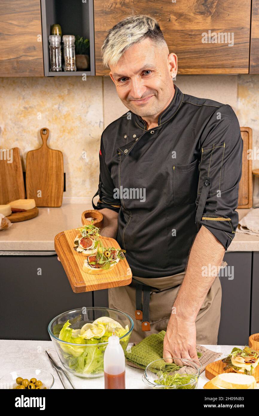 Le charmant chef tient la bruschetta italienne avec du fromage et des figues.Un chef élégant dans un uniforme dans sa cuisine.Beau chef de l'ethnie caucasienne pré Banque D'Images