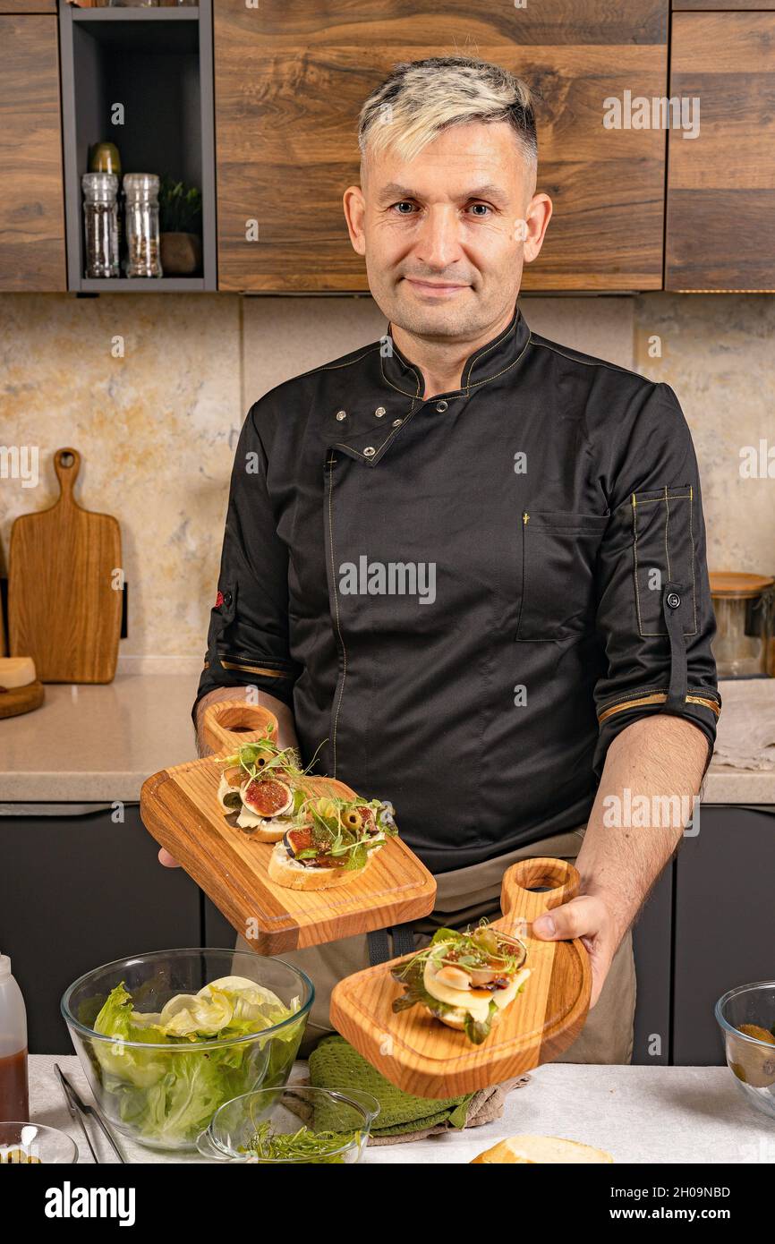 Le chef tient la bruschetta italienne avec du fromage et des figues.Un chef élégant dans une veste noire pose dans sa cuisine.Chef de l'ethnie caucasienne prepar Banque D'Images