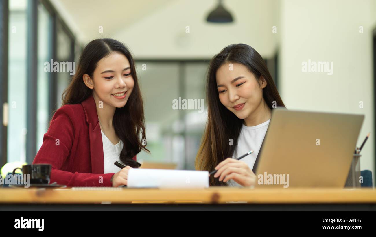 Deux jeunes femmes d'affaires souriantes qui travaillent et analysent ensemble les performances de l'impact social des entreprises sur le papier de rapport au bureau Banque D'Images