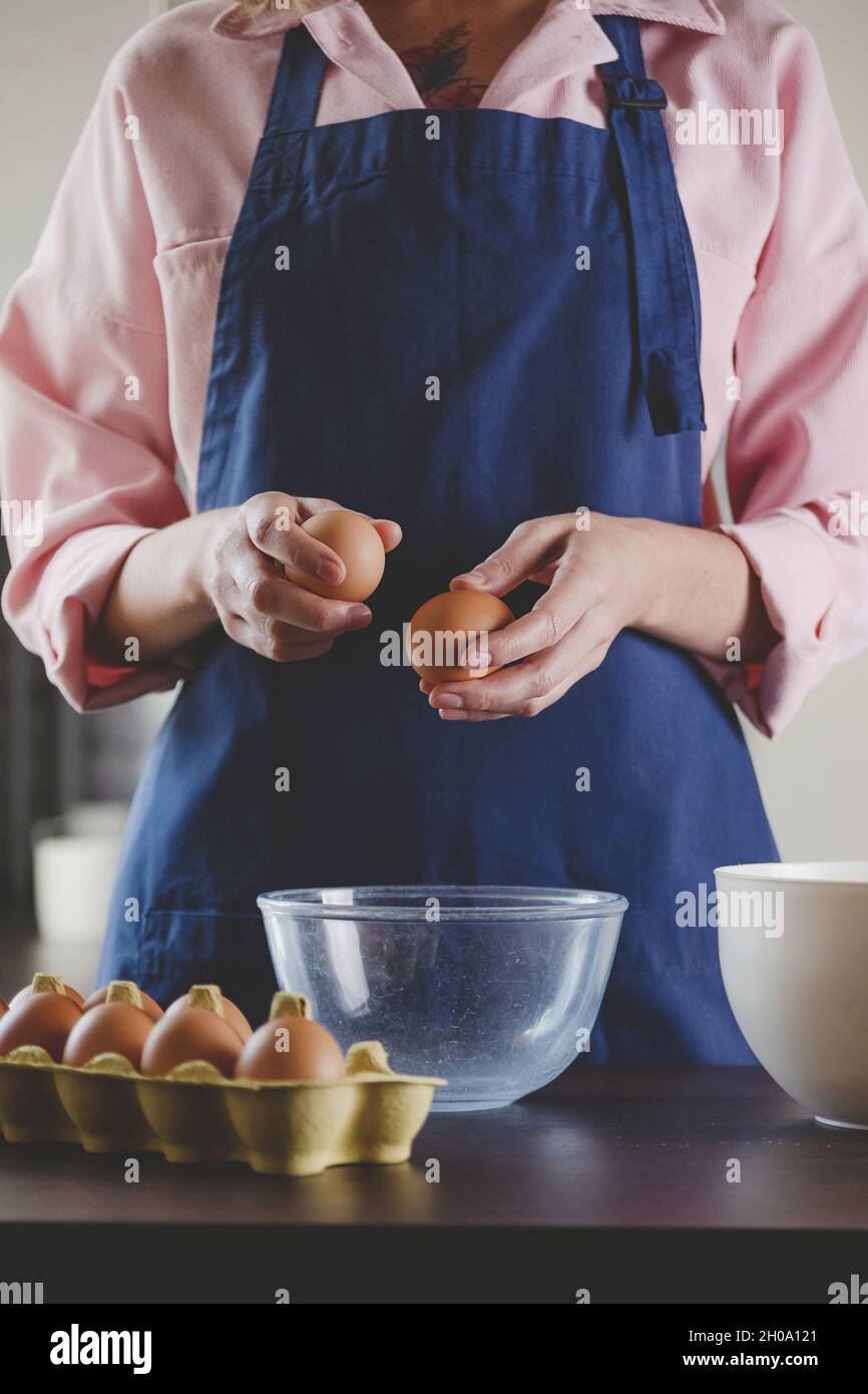 Femme boulangère dans un tablier bleu casse les oeufs pour la cuisson. Maison agréable esthétique de cuisine. Banque D'Images