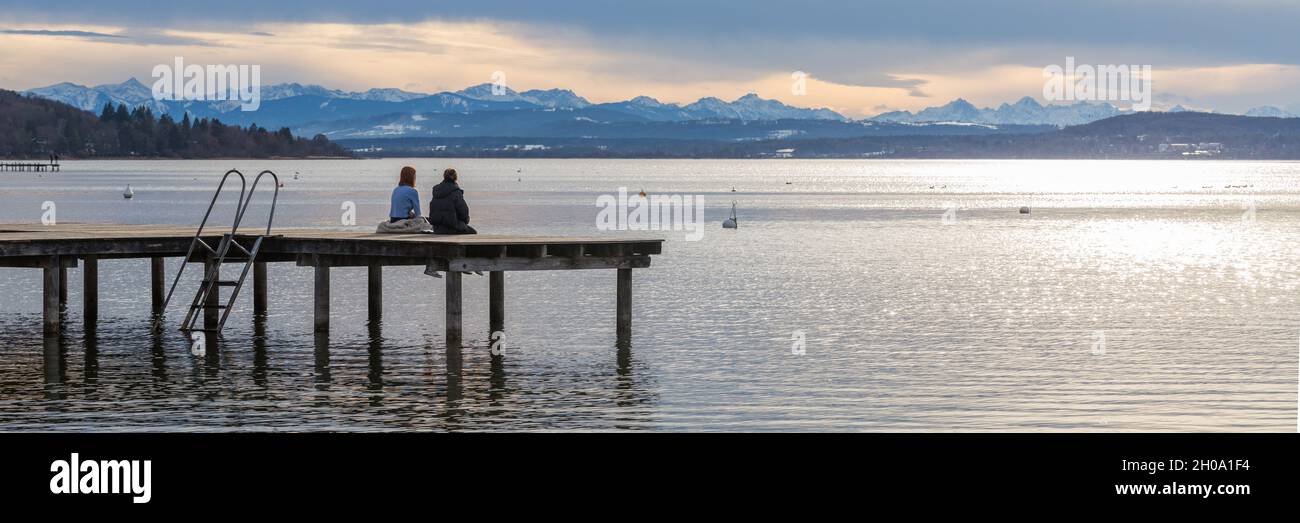 Herrsching, Allemagne - 22 janv. 2021: Panorama avec deux personnes assises sur un quai en bois au bord d'un lac (Ammersee).Montagnes à l'horizon. Banque D'Images
