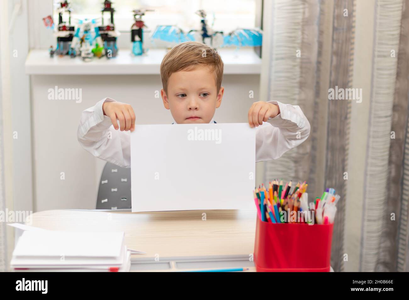 Un jeune garçon de première année dans un uniforme d'école à la maison tandis qu'isolé à son bureau tient une feuille de papier vierge. Mise au point sélective. Portrait Banque D'Images