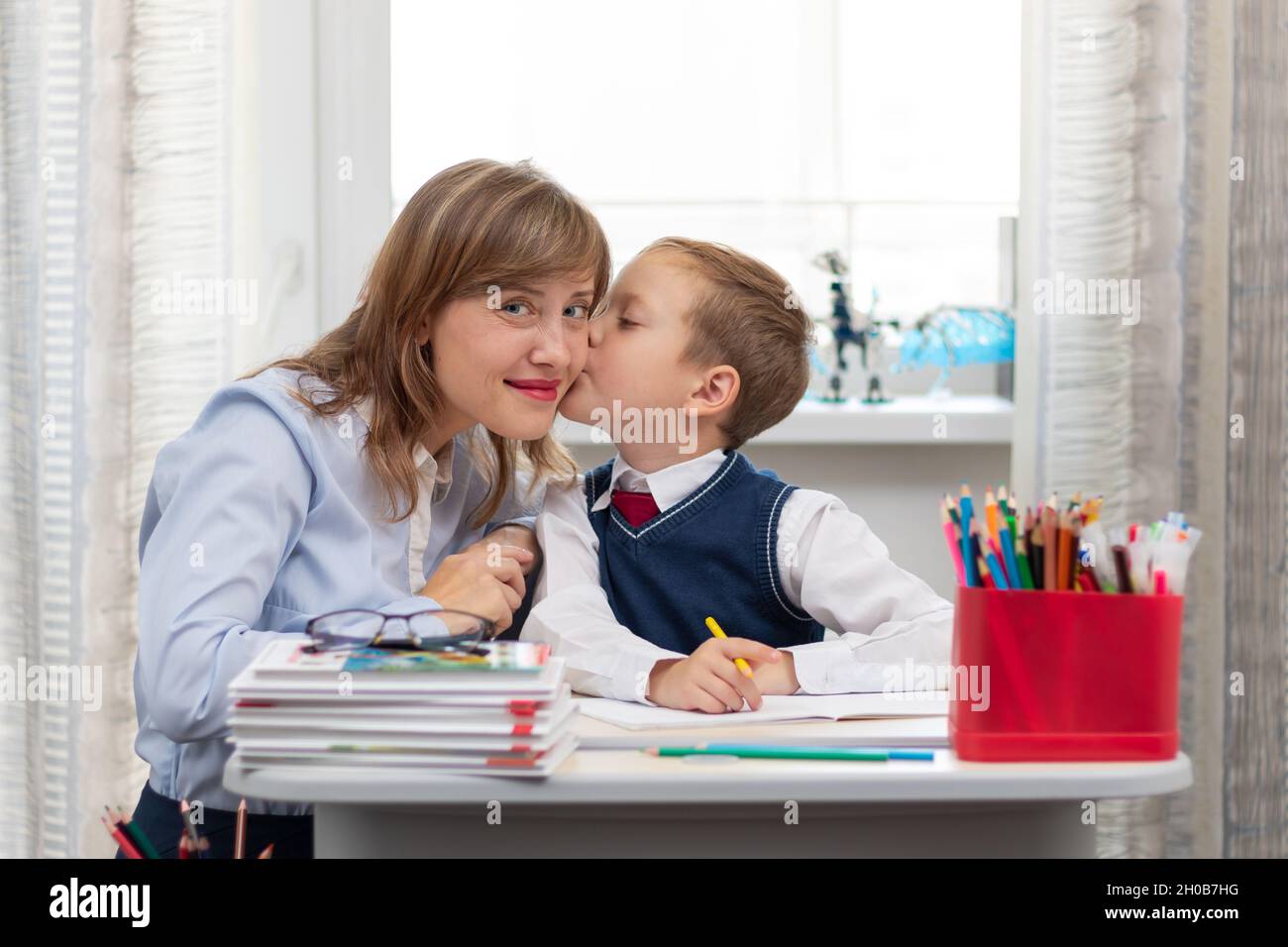 Une belle jeune mère avec son fils fait ses devoirs à un bureau à la maison contre un arrière-plan lumineux d'une fenêtre pendant une quarantaine pandémique. Sélection Banque D'Images