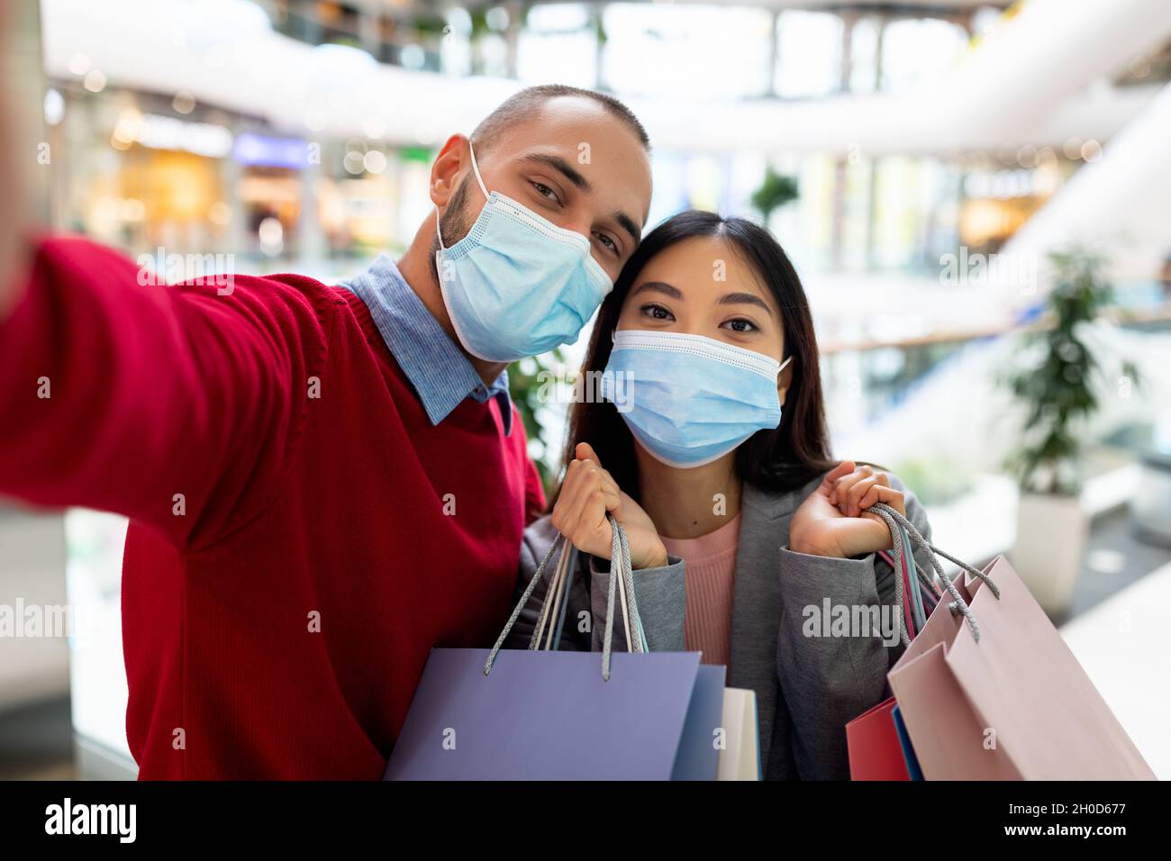 Jeune couple multiracial dans des masques de visage prenant le selfie ensemble tout en faisant du shopping dans un immense centre commercial Banque D'Images