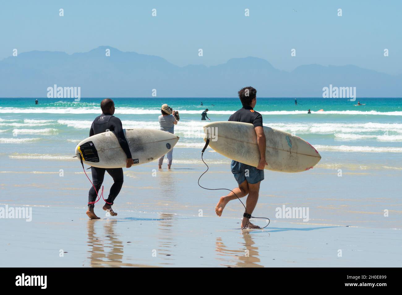 Les surfeurs courent dans la mer et les vagues à la plage de Muizenberg, au Cap occidental, en Afrique du Sud, tenant des planches de surf sous leurs bras Banque D'Images