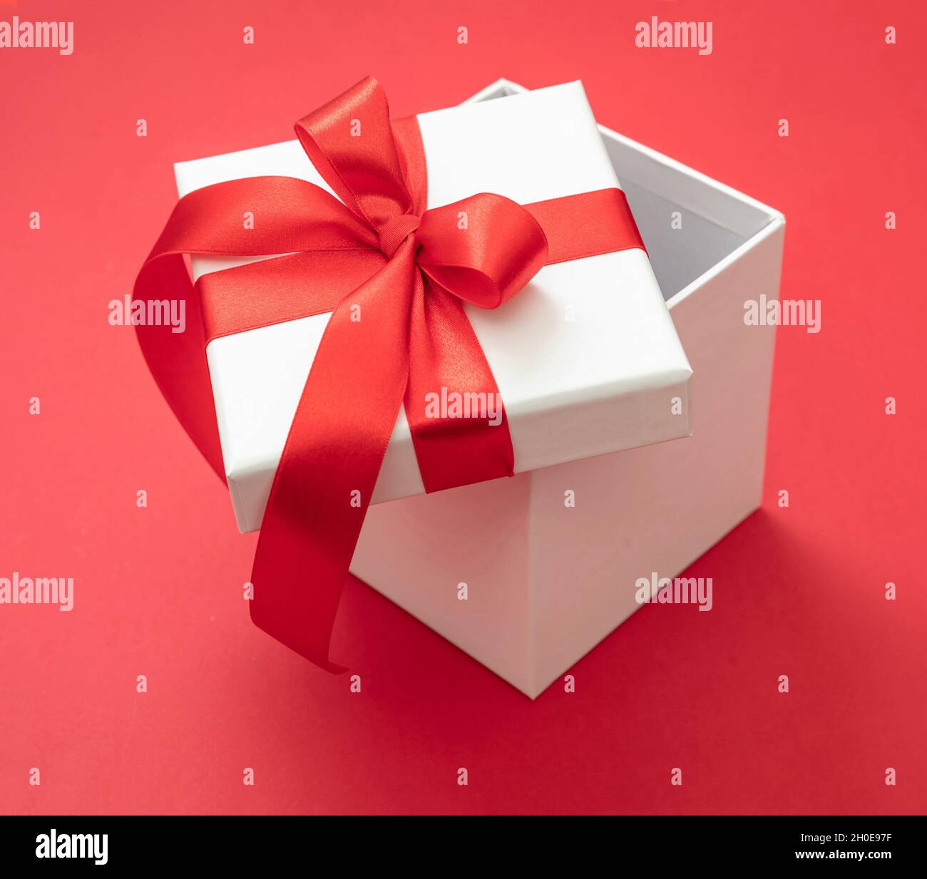 Boîte cadeau de Noël blanche avec ruban rouge noeud ouvert sur fond rouge, surprise de la Saint-Valentin, cadeau de vacances du nouvel an, décoration en satin curly. Banque D'Images