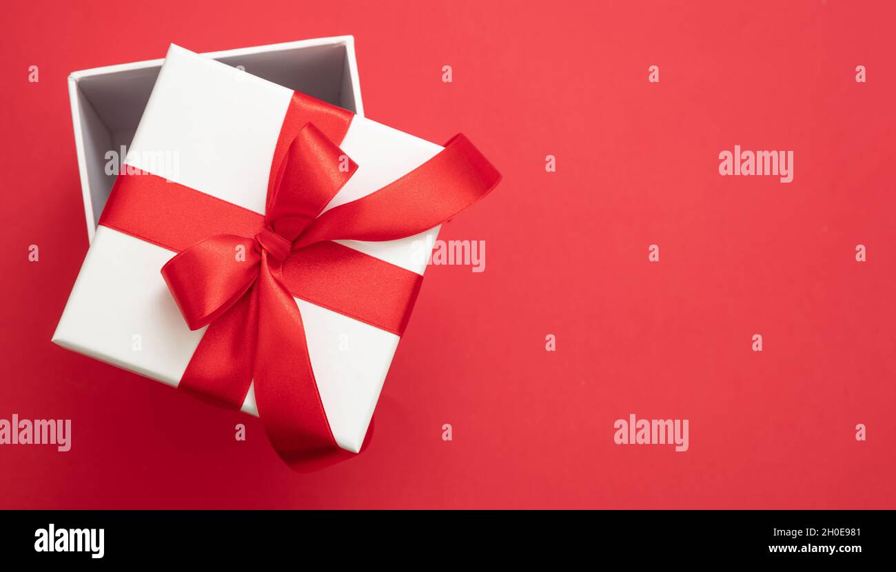 Boîte cadeau de Noël blanc avec ruban rouge noeud ouvert sur fond rouge couleur, surprise de la Saint-Valentin, cadeau de vacances de nouvel an, décoration de curly en satin, haut v Banque D'Images