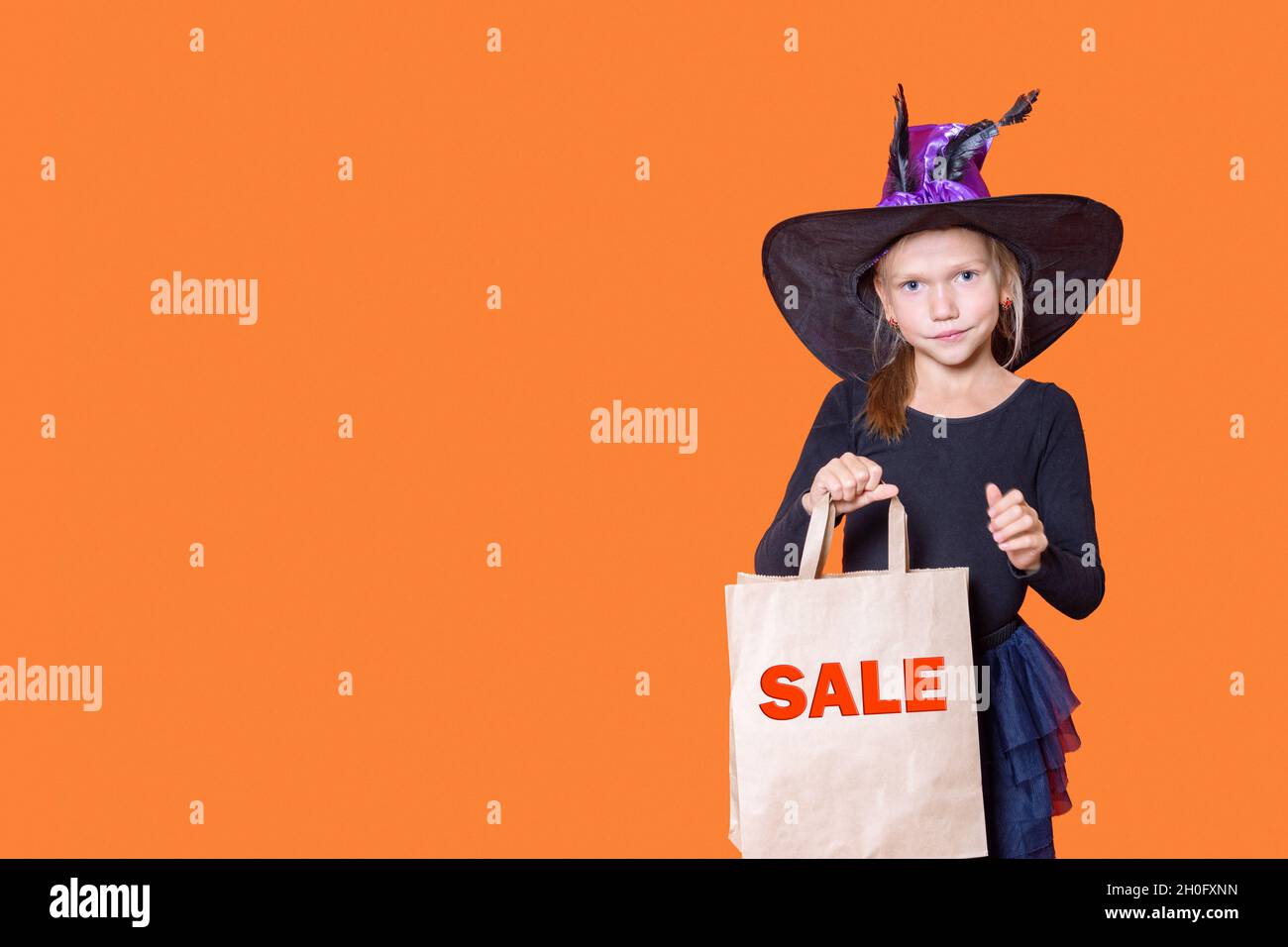 Une belle fille souriante dans une robe noire et un chapeau de sorcière tient un sac de papier d'artisanat avec les mots VENDUS sur un fond orange.Conc. Shopping Halloween Banque D'Images