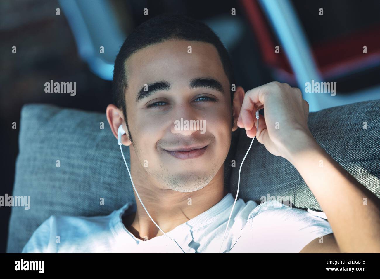 Jeune homme noir souriant avec un chaume non rasé écoutant de la musique sur des bouchons d'oreille en gros plan pendant qu'il se détend sur un canapé au soleil Banque D'Images