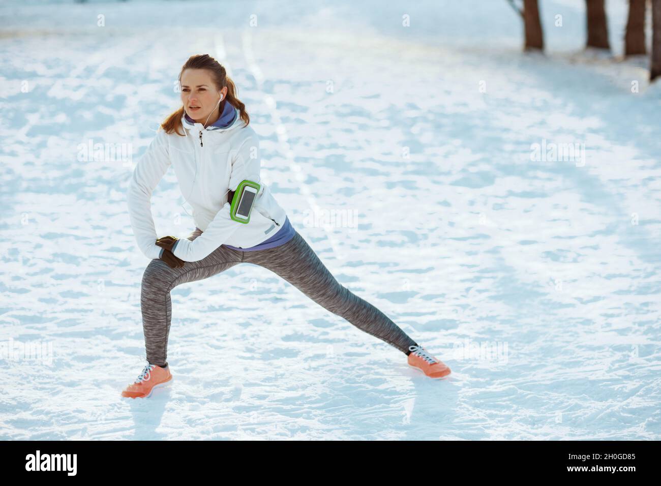 Portrait complet de la femme active en veste blanche s'étendant à l'extérieur dans le parc de la ville en hiver. Banque D'Images