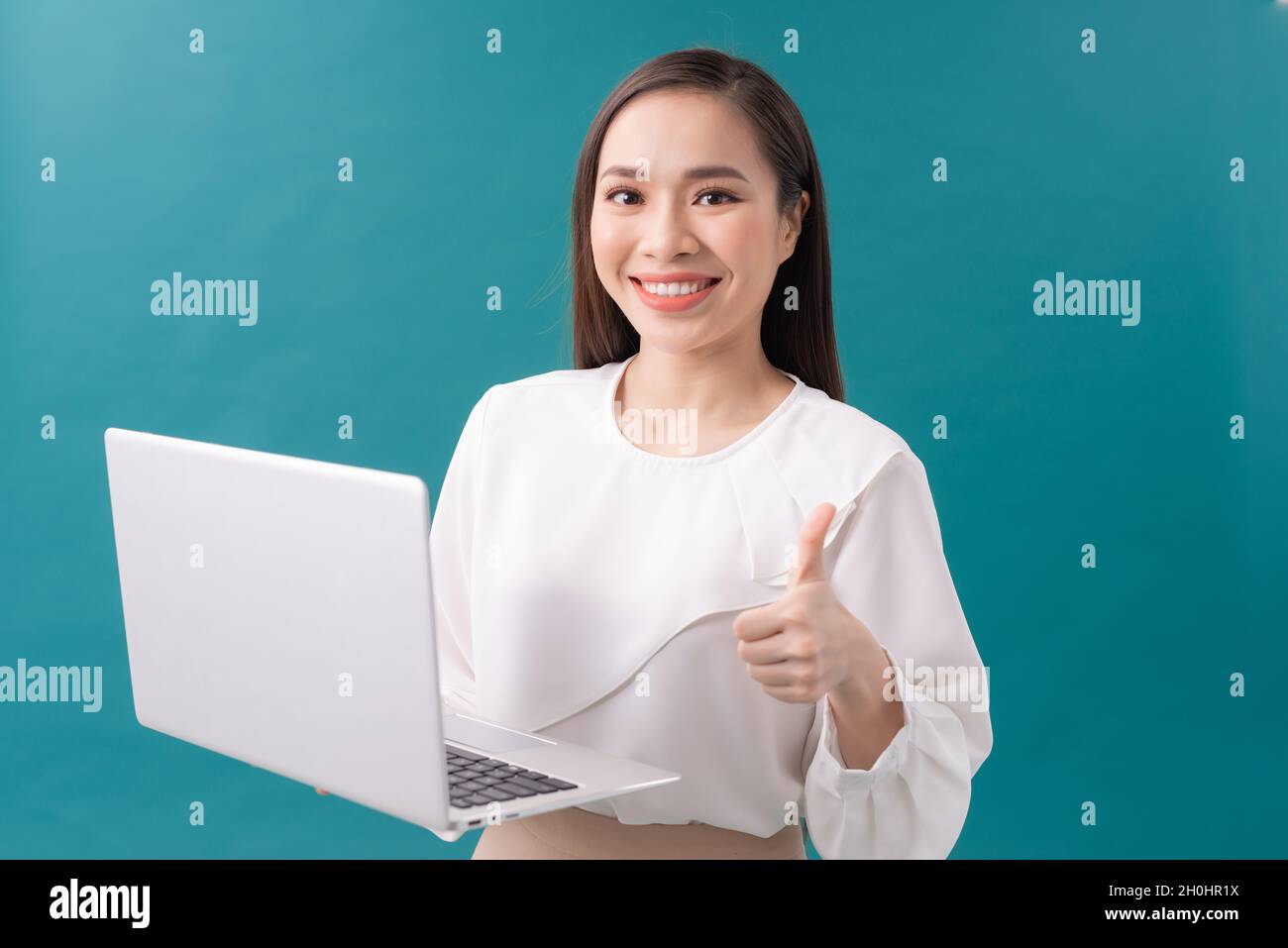 Tenue d'une femme d'affaires asiatique tenant un ordinateur portable et montrant le pouce vers le haut tout en regardant la caméra sur fond bleu Banque D'Images