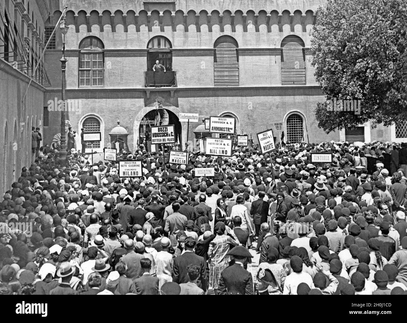 "Les travailleurs de l'industrie textile autour de Milan écoutent le chef de gouvernement italien Benito Mussolini (sur le balcon) lors d'un discours en 1930.Les travailleurs sont originaires de : ''Industria Sete Cucirine'' de Milan et de Novate, manufacture ''Cucirini Cantoni Coats' de Maglio, société ''Bozzi Vidossich and C''' de Milan et Masat.[traduction automatique]' Banque D'Images