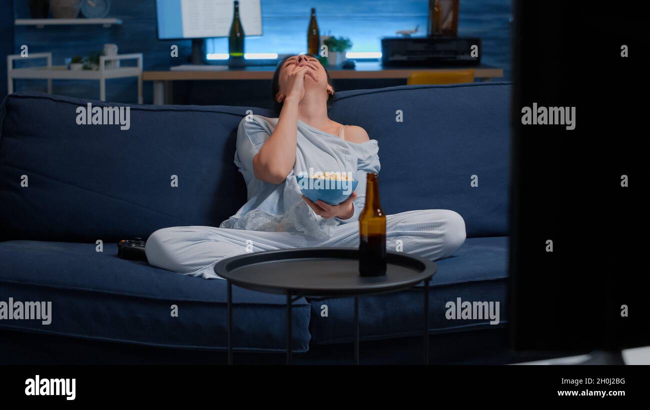 Une jeune femme heureuse se détendant sur un canapé confortable, mangeant du pop-corn rire en regardant un film amusant en ligne, montrer des programmes divertissants.Famele passer du temps libre le week-end seul à la maison vêtu de pyjama Banque D'Images