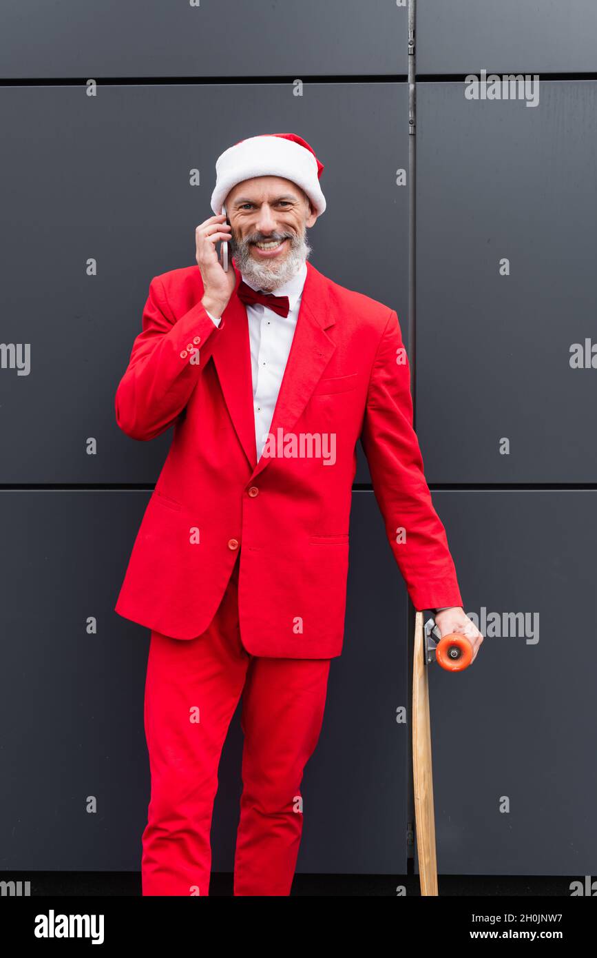 heureux homme mature dans le chapeau de santa parlant sur le téléphone portable tout en se tenant avec le skateboard près du mur gris Banque D'Images