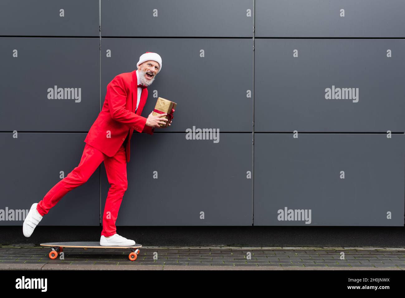 heureux homme mature dans le chapeau de santa tenant présent et de l'équitation sur le skateboard près du mur gris Banque D'Images