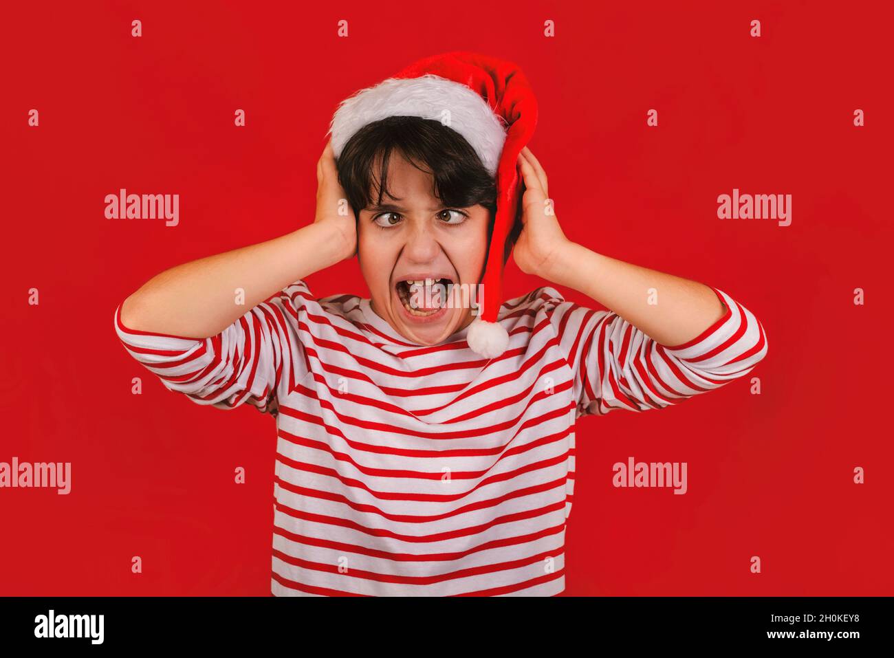 Enfant en colère portant le chapeau du Père Noël avec ses mains sur la tête sur fond rouge Banque D'Images