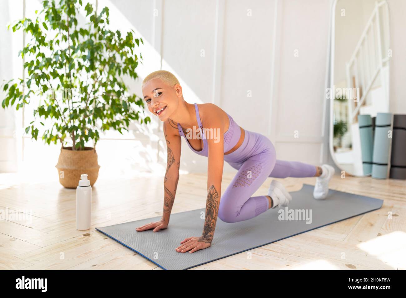 Bonne dame avec une coupe courte de cheveux faisant des exercices de alpinistes de corps de croix, s'exerçant sur le tapis de yoga dans l'appartement lumineux Banque D'Images