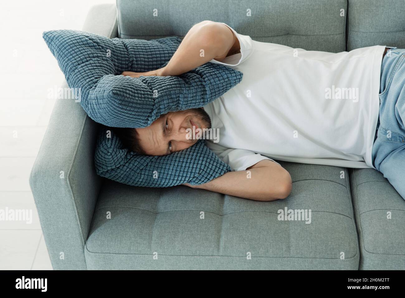 L'homme agacé couvre les oreilles avec des oreillers pour éviter le bruit de voisin en se nappant sur le canapé essayer de dormir Banque D'Images