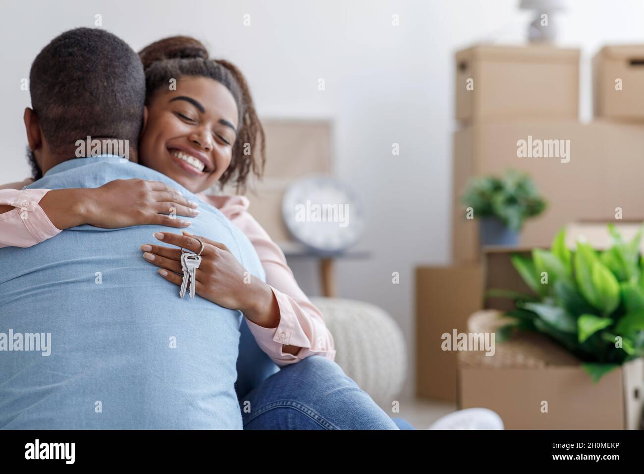 Une jeune famille noire satisfaite se serrer, tenir la clé et profiter d'acheter de l'immobilier dans la chambre avec des boîtes en carton et des plantes Banque D'Images