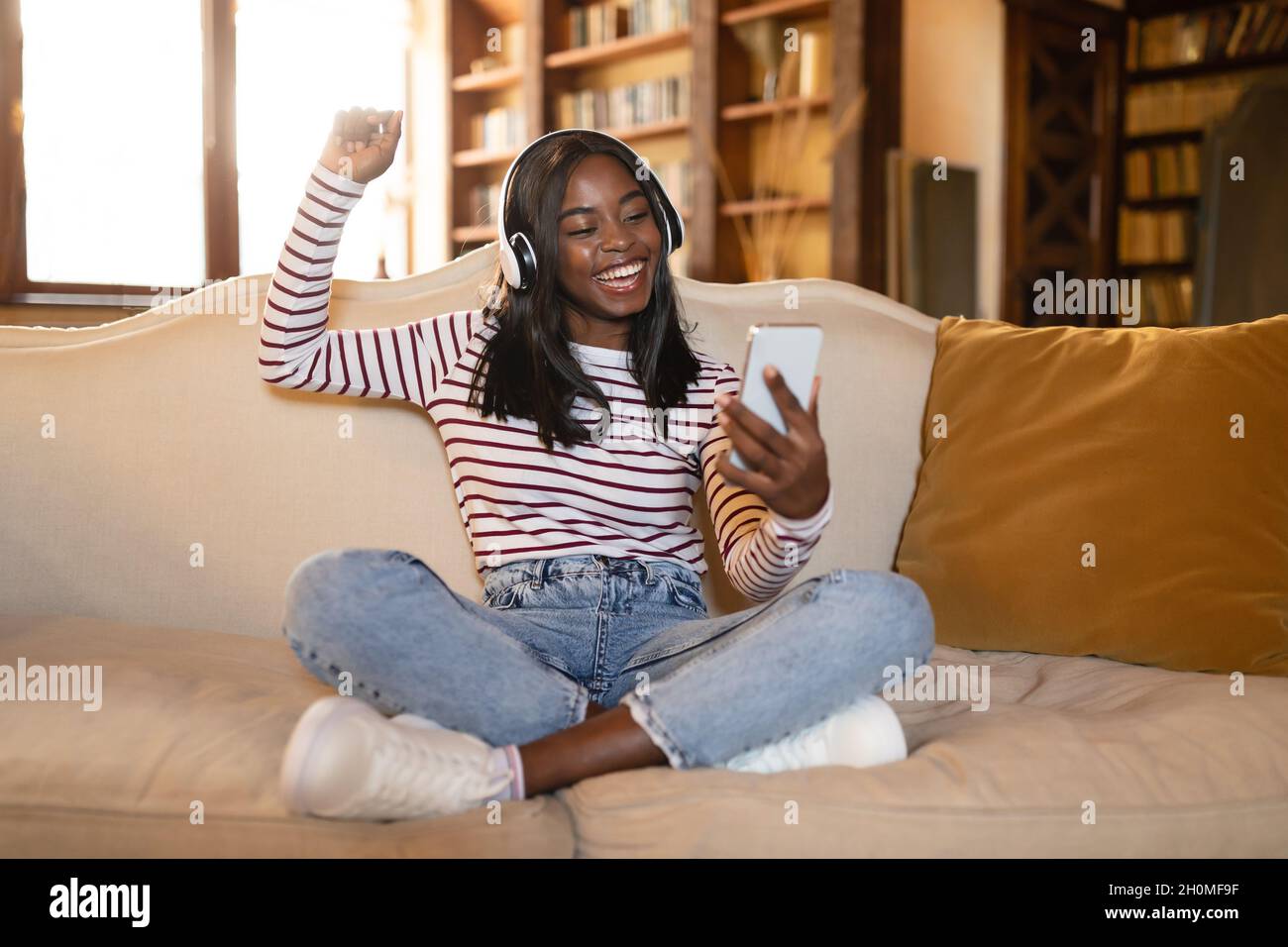 Jeune femme noire positive avec casque et téléphone mobile dansant tout en étant assise sur le canapé et en écoutant de la musique à la maison Banque D'Images