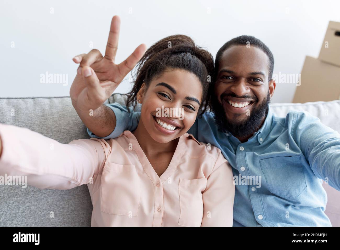 Un jeune couple noir souriant se repose sur le canapé, l'homme montre le signe de la paix, fait le selfie dans la chambre Banque D'Images