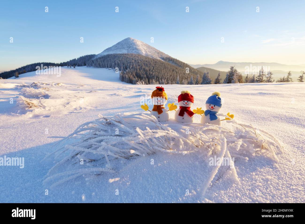 Trois petits bonhommes de neige en chapeau et foulard sur une pelouse enneigée.Magnifique coucher de soleil en hiver.Paysage avec hautes montagnes.Joyeux noël et joyeux nouveau Banque D'Images