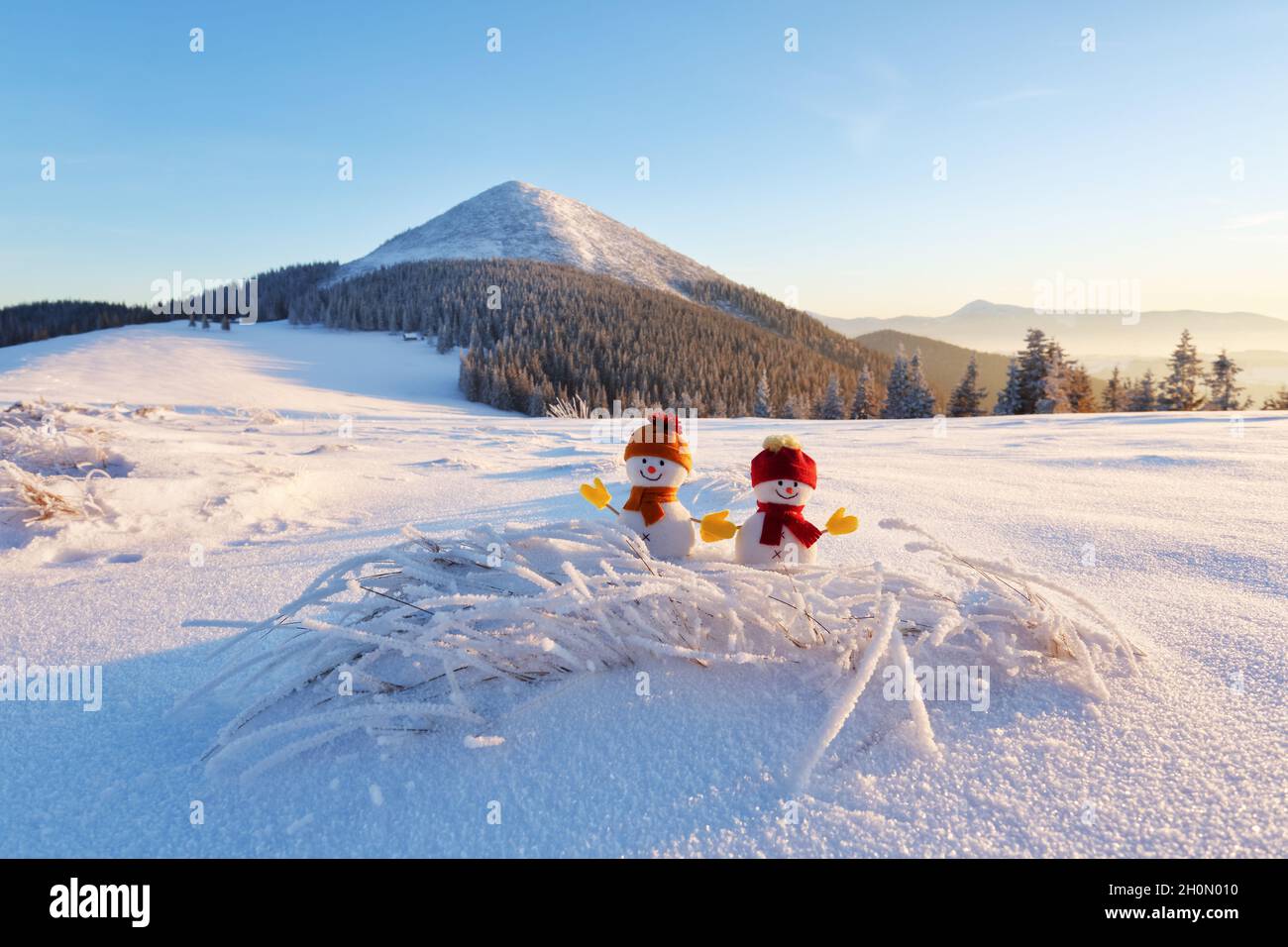 Couple de deux petits bonshommes de neige en chapeau rouge et foulard sur un terrain enneigé. Magnifique coucher de soleil en hiver. Paysage avec hautes montagnes. Joyeux noël et Banque D'Images