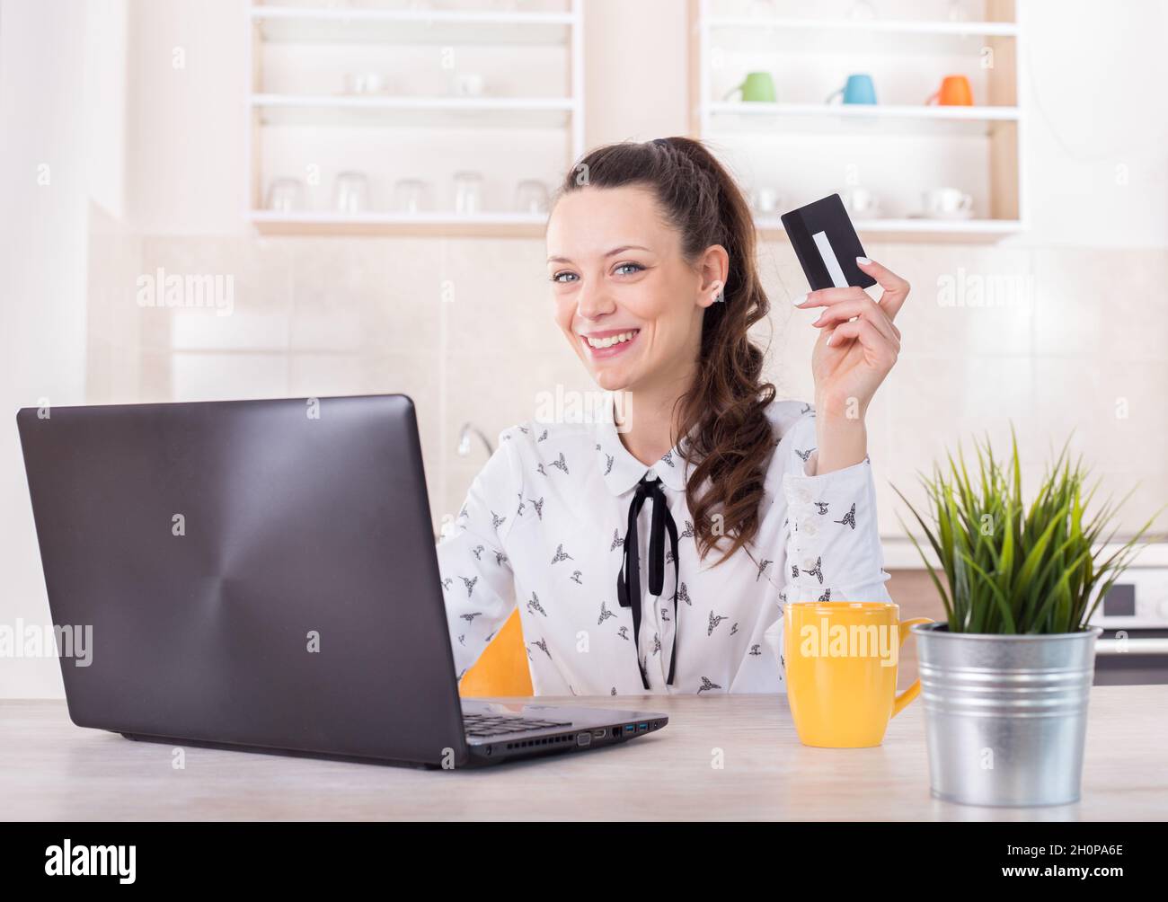 Bonne jeune femme assise à la table de cuisine avec un ordinateur portable et tenant une carte de crédit.Achat en ligne et e-business concept Banque D'Images