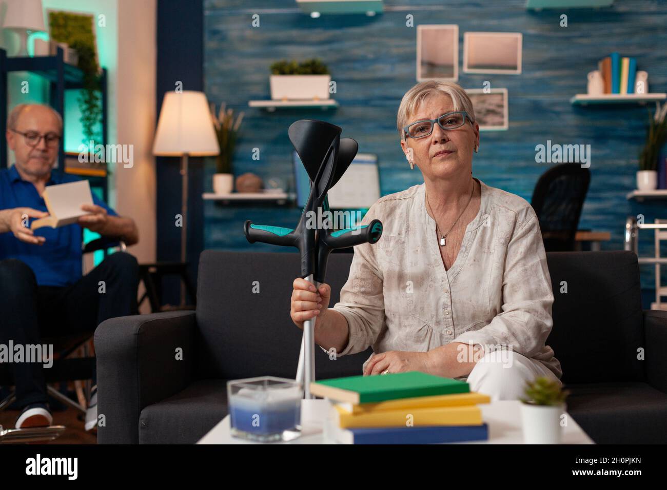 Femme âgée tenant des béquilles et regardant la caméra dans l'espace intérieur.Personne âgée handicapée assise sur un canapé alors que l'homme à la retraite lit en fauteuil roulant dans le fond de la salle de séjour Banque D'Images