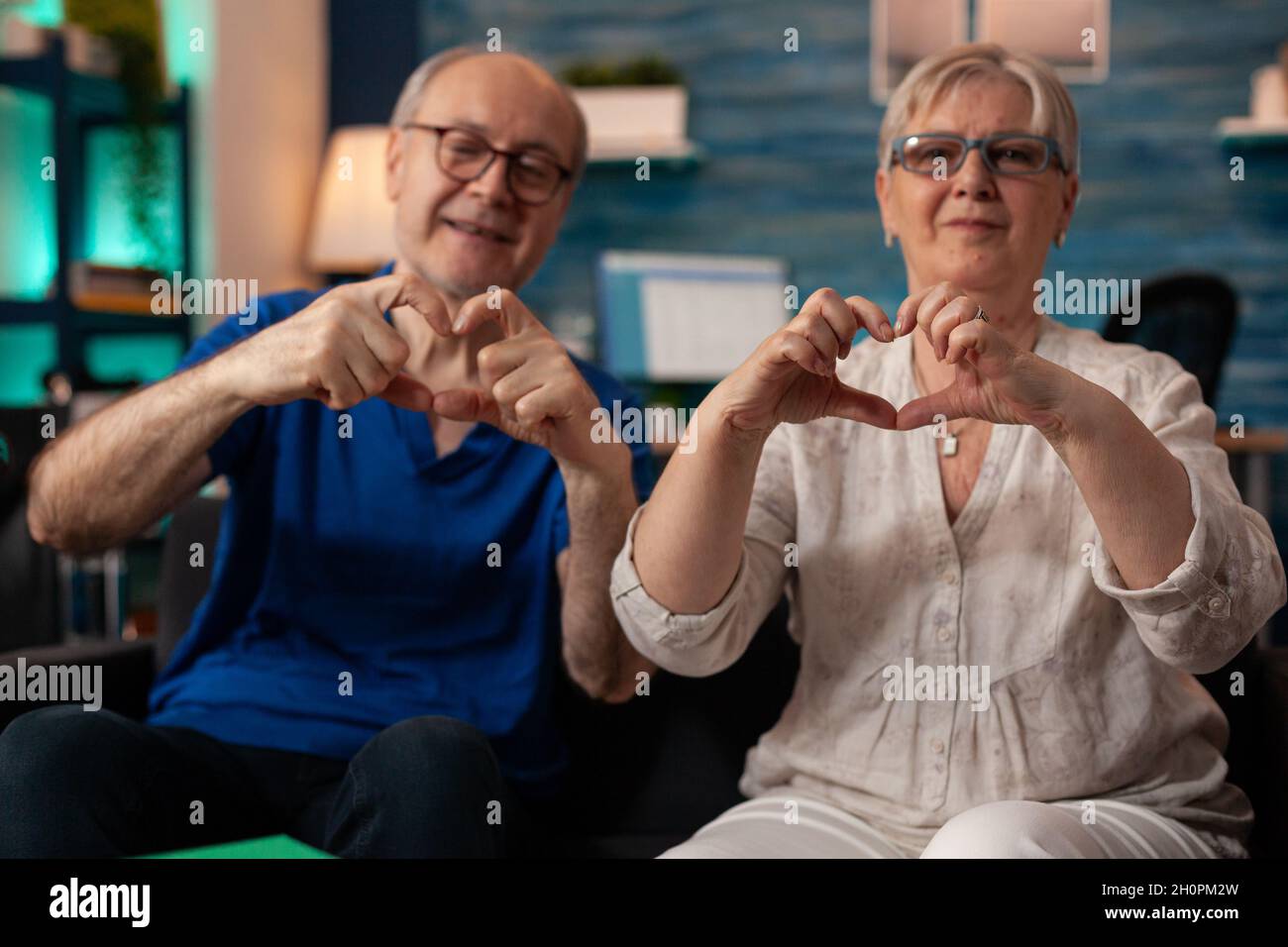 Un vieux couple marié faisant le symbole du cœur avec les mains tout en regardant la caméra dans le salon.Mari et femme à la retraite créant un geste romantique d'amour assis sur le canapé à la maison.Relation senior Banque D'Images